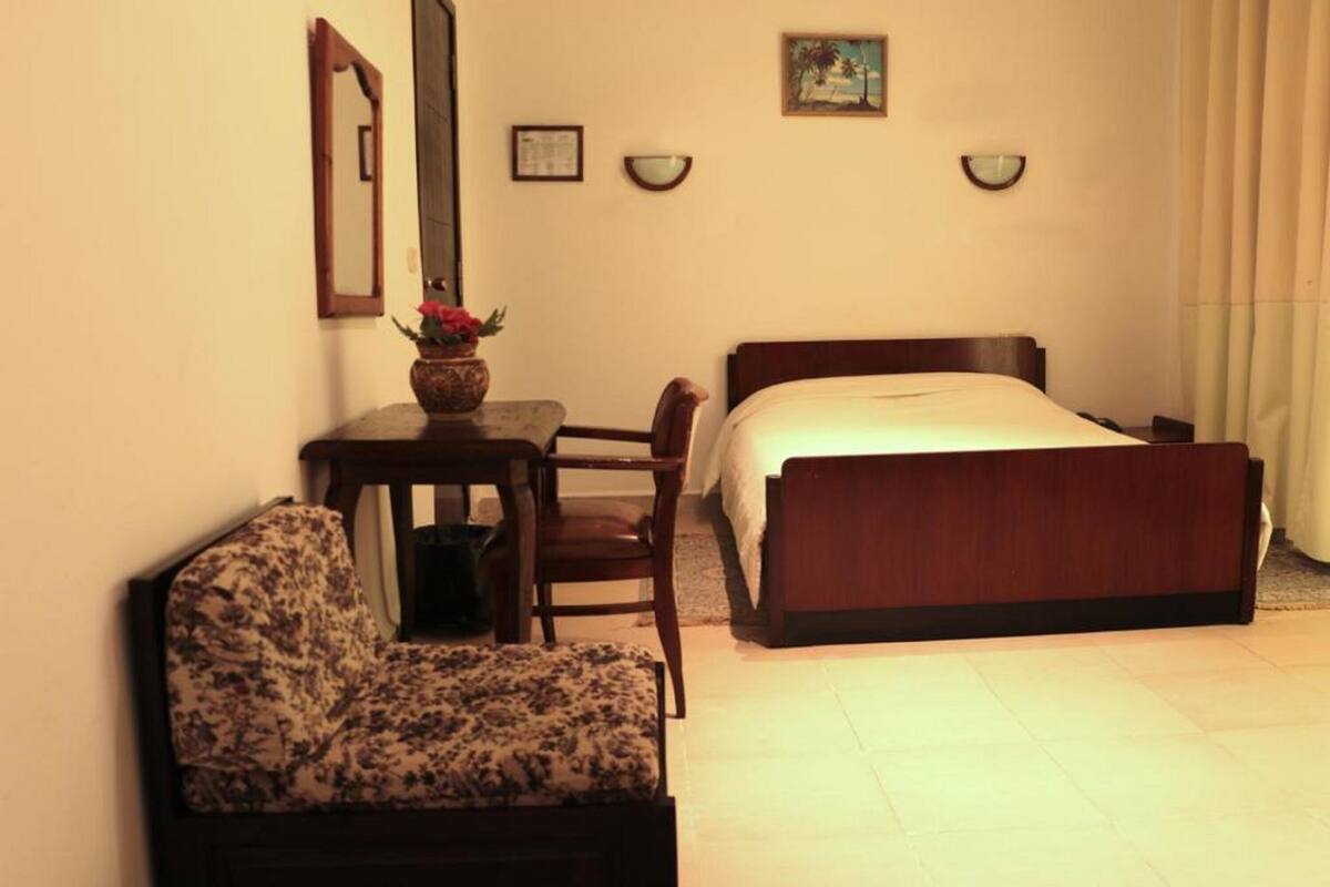 A bedroom with a bed, a chair, a small table with a vase, and a mirror on the wall.