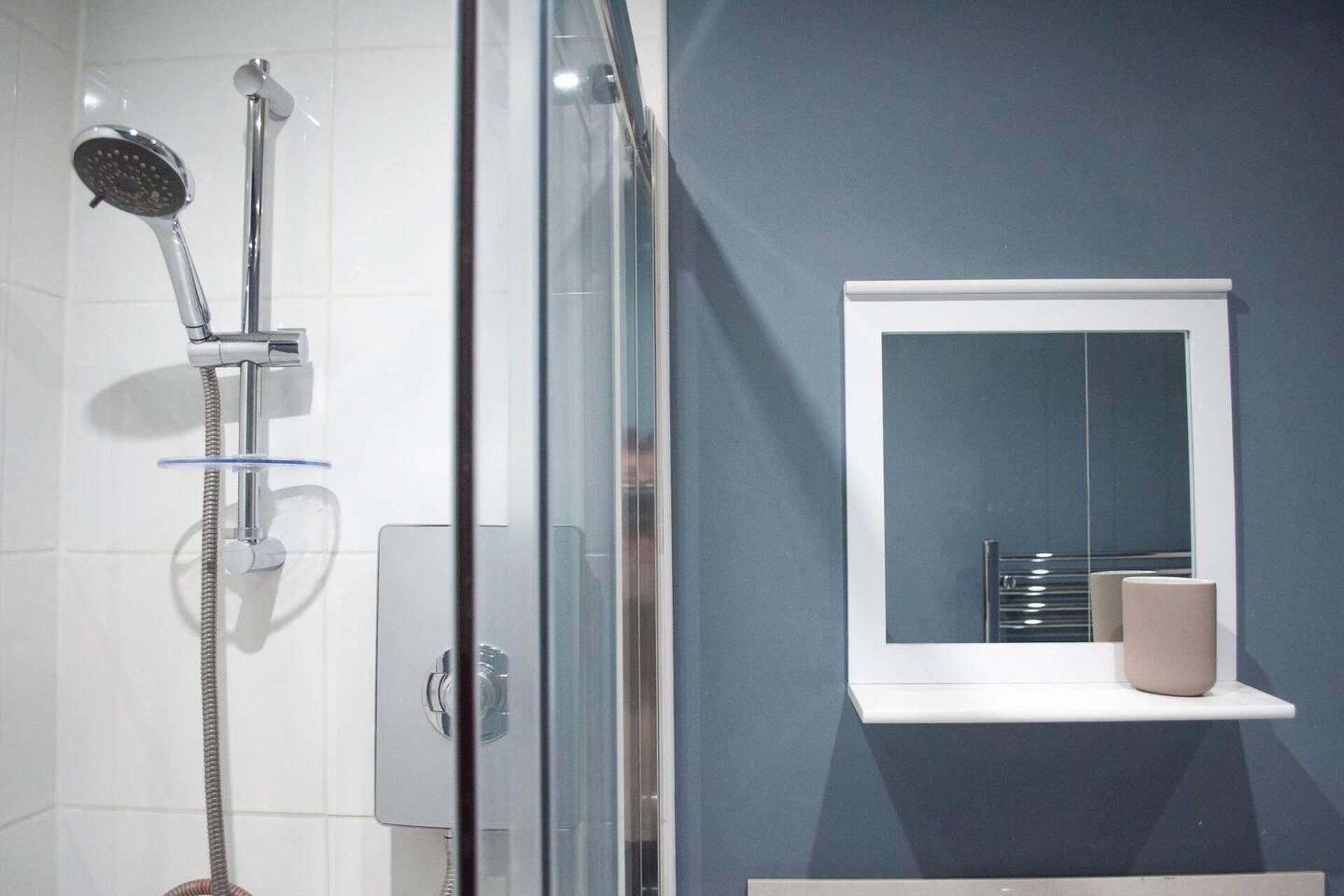A modern bathroom with a glass-enclosed shower, a white shelf with a cup, and a blue wall.