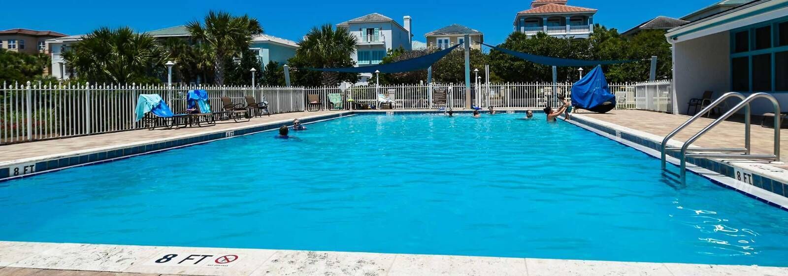 Una piscina con indicador de profundidad de 2.4 metros, rodeada por una valla blanca, con sillas de playa y sombrillas, y edificios al fondo.