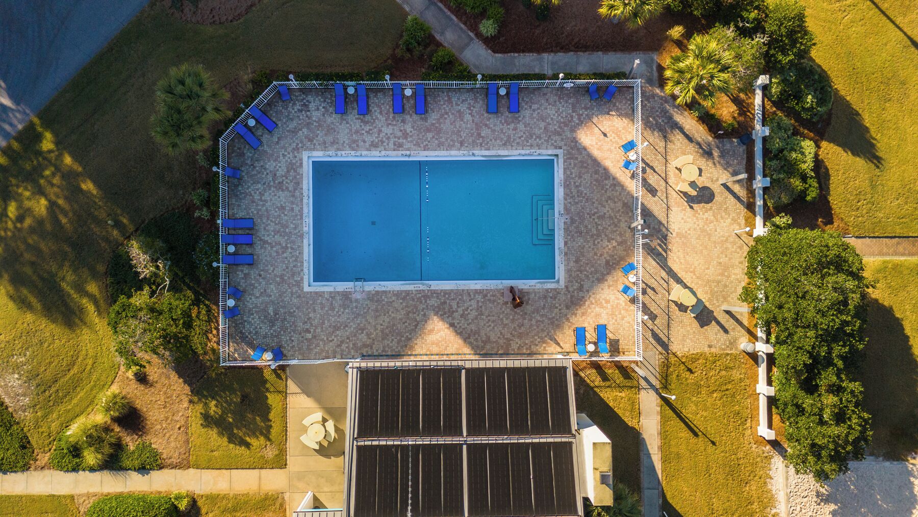 An aerial view of a rectangular swimming pool surrounded by a tiled deck with blue lounge chairs and a solar panel array.