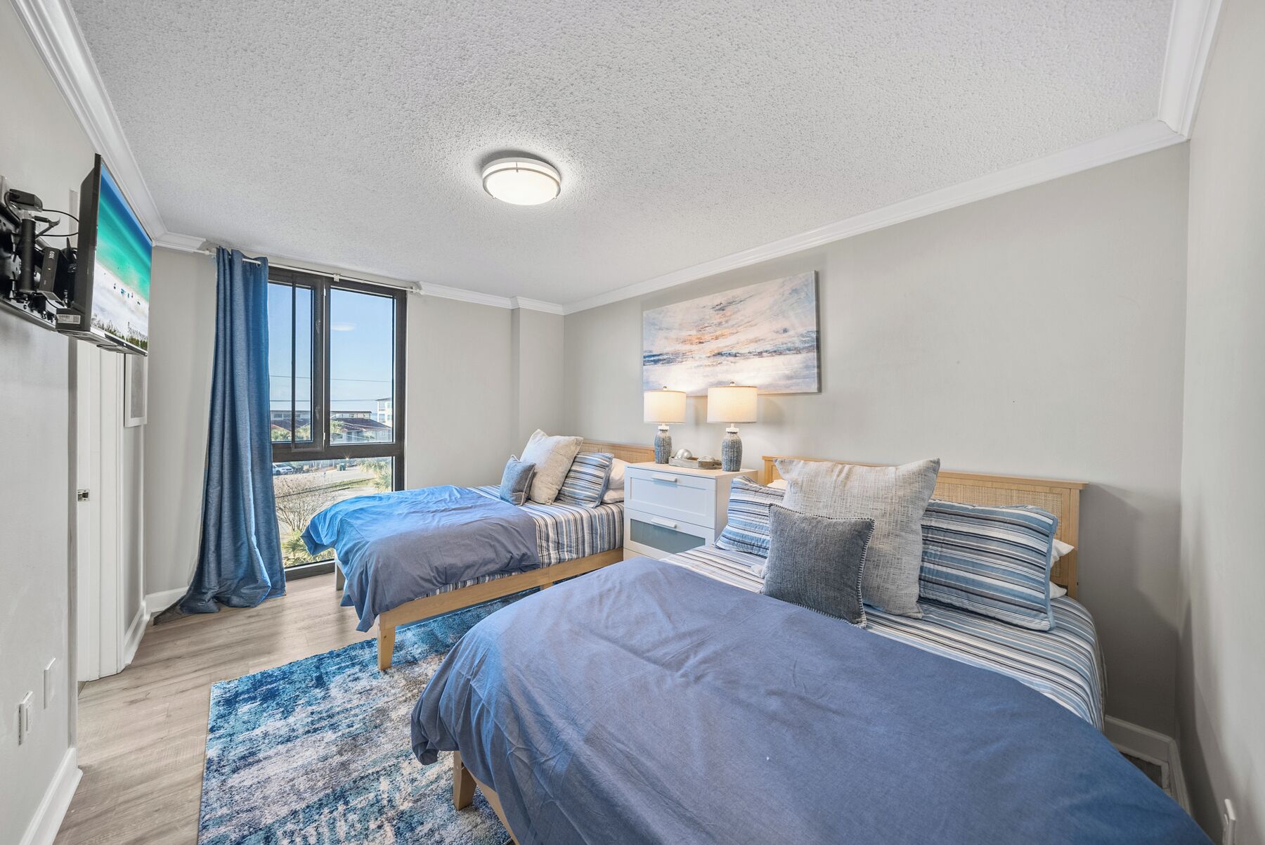 A bedroom with two beds, a nightstand, a lamp, a painting, and a view of the outdoors.