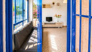 Ein Zimmer mit einer Couch, einem Fernseher, einer kleinen Kommode und einem Fenster mit blauen Rahmen.