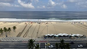 A beach with a road in the foreground, palm trees, and a clear view of the ocean. - Frente Mar Encantador (Rio de Janeiro)