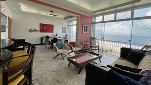 A spacious living area with a high ceiling, a ceiling fan, a dining table with chairs, a sofa, and a view of the sea through large windows. - Frente Mar Encantador (Rio de Janeiro)