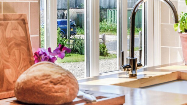 Una cocina con una hogaza de pan sobre una tabla de cortar, un lavamanos y una ventana con vistas a un jardín.