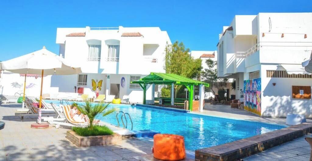 Una piscina con sillas de descanso, un área de estar cubierta y un edificio de paredes blancas y balcones.