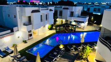 A rooftop pool area with lounge chairs and umbrellas, surrounded by white buildings with balconies.