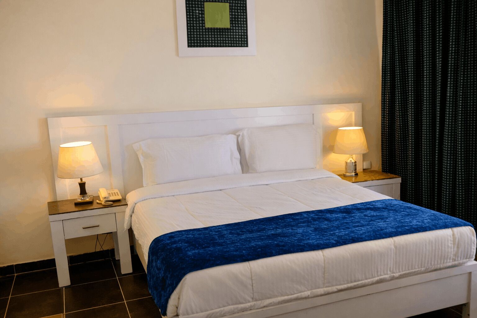A hotel room with a bed, two bedside tables, a nightstand, a lamp, a phone, and a wall-mounted light fixture.