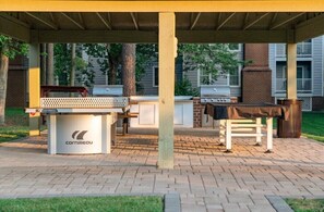Ein Außenbereich mit Grill, Tisch und Ziegelterrasse, umgeben von Bäumen.