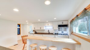 A modern kitchen with white cabinets, a wooden island with stools, a built-in microwave, and a window with a view. - Walk to Beach! Sodus Point Escape w/ Private Yard (Sodus Point)
