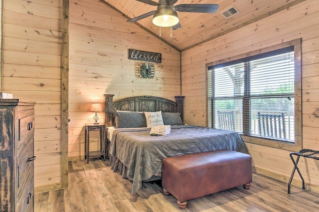 A cozy bedroom with a wooden ceiling, a bed with a headboard, a bench, a nightstand, and a window with blinds.