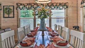 Un espace repas avec une longue table dressée pour un repas, entourée de chaises, un vase central rempli de fleurs et un luminaire décoratif au plafond.