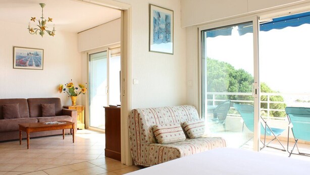 A living room with a sofa, a coffee table, a patterned armchair, and a bed. There is a balcony with chairs and a view of greenery. - Apartment for 6 in a standing residence with swimming pool (CAVALAIRE sur mer)