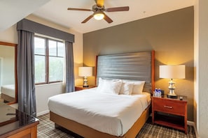 Quarto de hotel com cama, duas mesas de cabeceira, ventoinha no teto, janela com cortinas e espelho.