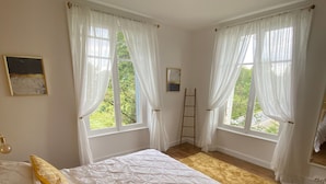 Ein Schlafzimmer mit einem großen Bett, zwei Fenstern mit weißen Vorhängen, einem Holzboden und einer an der Wand stehenden Leiter.