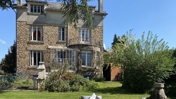 Ein Steinhaus mit großem Vorgarten, Sitzbereich im Freien mit Holzmöbeln und einem klaren, blauen Himmel.