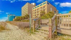 Een hotel aan het strand met een houten looppad naar de ingang, een helderblauwe lucht en palmbomen.