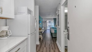 Een smalle gang met een kitchenette aan de linkerkant, een eetgedeelte met tafel en stoelen in het midden, en een bed met raam aan de rechterkant.