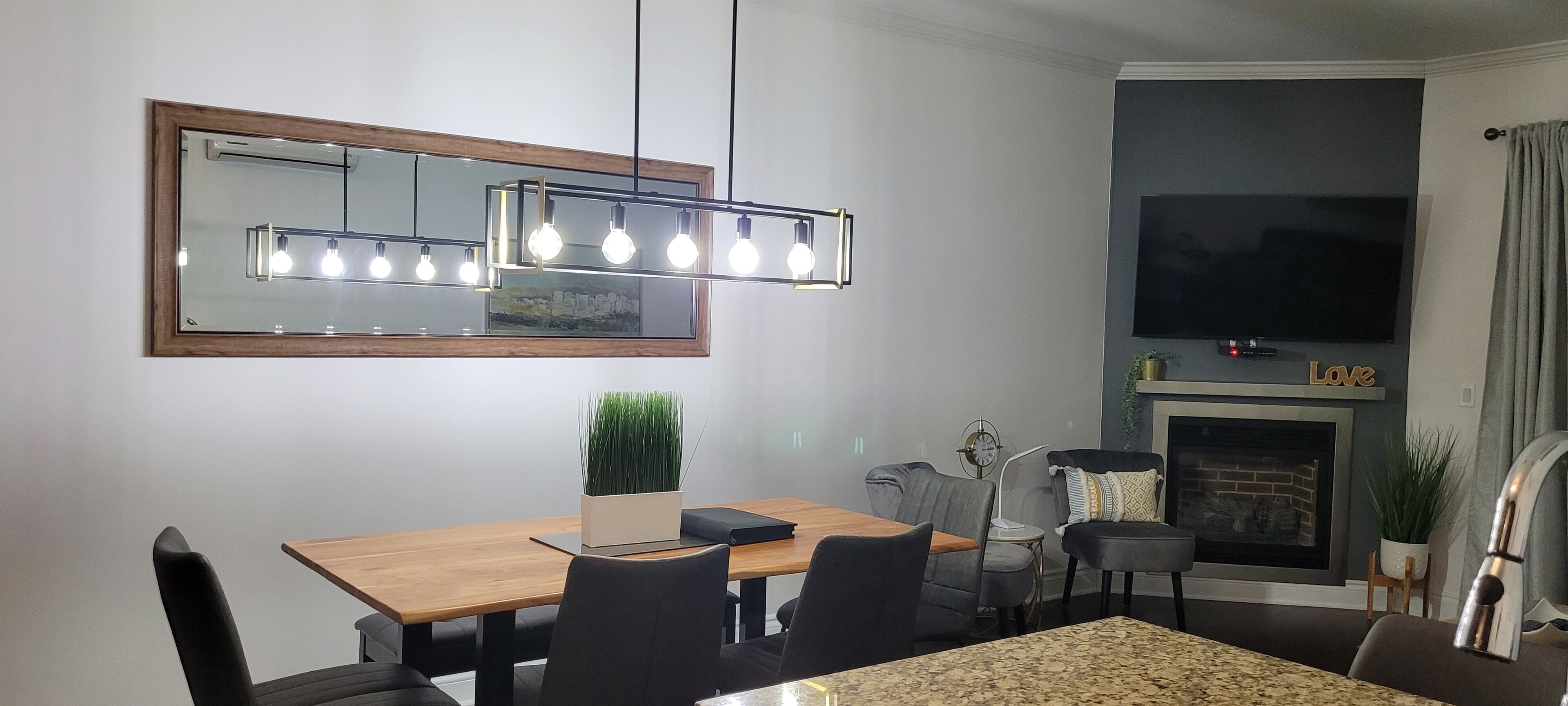A modern dining area with a wooden table, black chairs, a mirror with lights, a TV, and a fireplace.