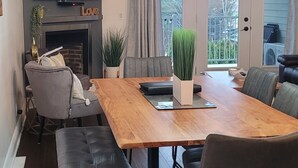 Un salon moderne avec une table à manger en bois, des chaises recouvertes d’un tissu gris, un téléviseur à écran plat fixé au-dessus d’une cheminée et une grande fenêtre agrémentée de rideaux.
