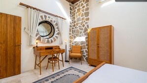 A bedroom with a bed, a wooden desk, two chairs, a round mirror, a stone wall, and a wooden wardrobe. - Tropical villa style • Sleeps six • Berawa (Kecamatan Kuta Utara)