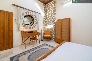 A bedroom with a bed, a wooden desk, two chairs, a round mirror, a stone wall, and a wooden wardrobe. - Tropical villa style • Sleeps six • Berawa (Kecamatan Kuta Utara)