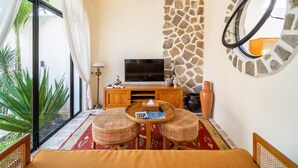 A room with a TV, wicker furniture, a stone wall, and a large window. - Tropical villa style • Sleeps six • Berawa (Kecamatan Kuta Utara)