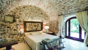 Slaapkamer met stenen muren, een bed, nachtkastje, lamp, raam met gordijnen en uitzicht op groen buiten.