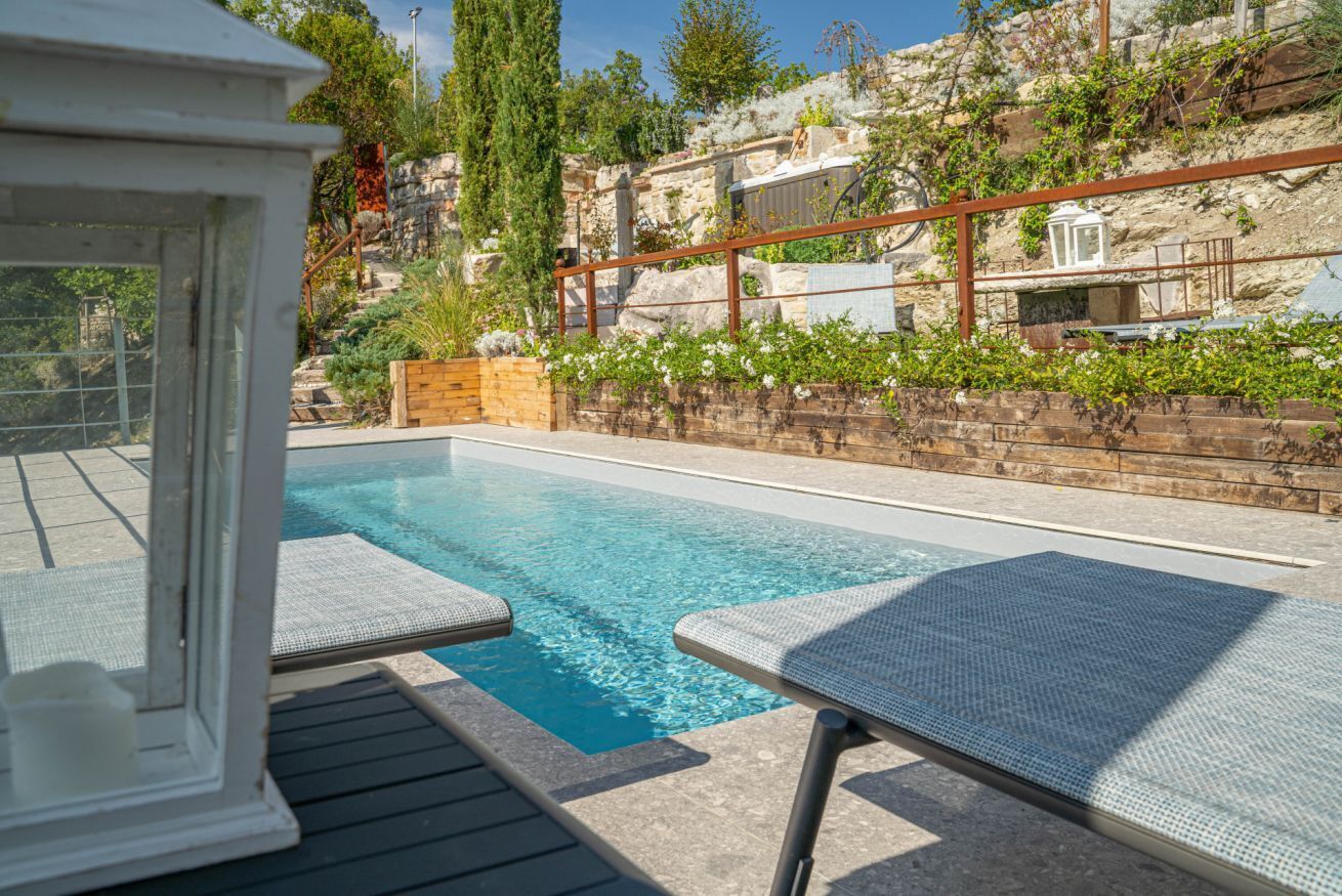 Une piscine entourée de chaises longues, d’une terrasse en bois et d’un mur de pierre agrémenté de verdure.