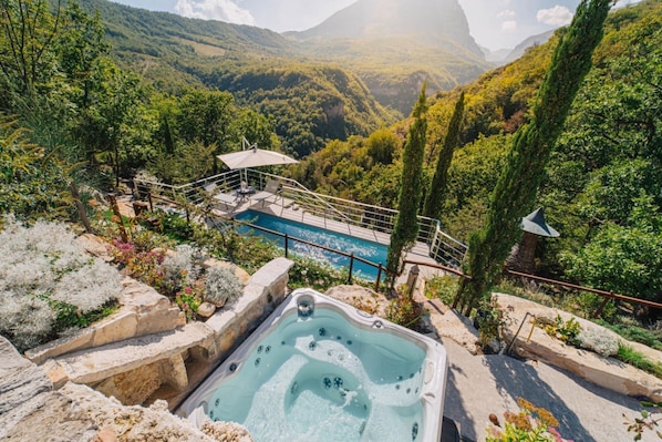 A hot tub surrounded by a pool, with a mountainous backdrop and lush greenery.