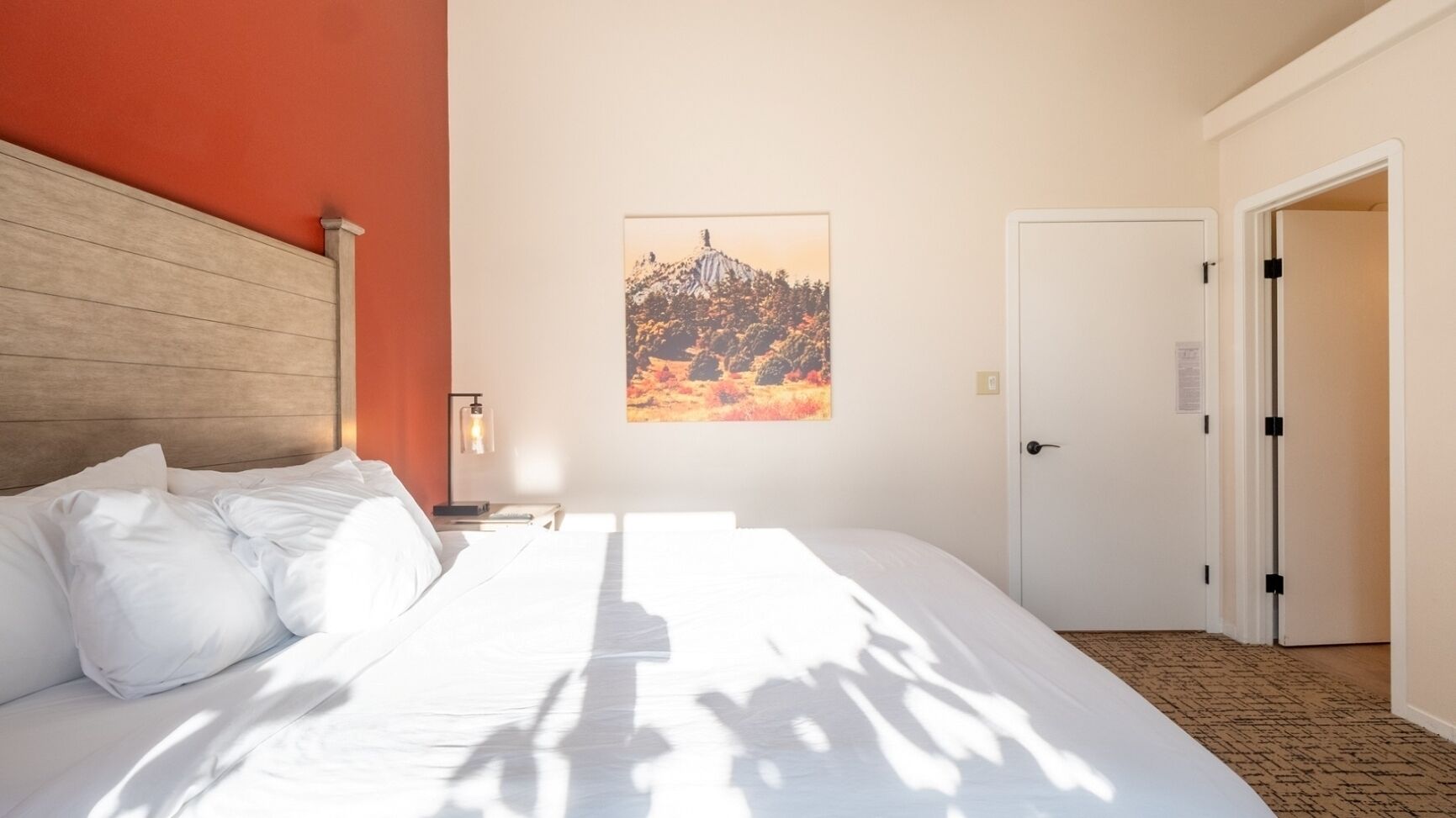 Une chambre d’hôtel avec un grand lit, une table de chevet avec une lampe, un tableau au mur et une porte sur le côté droit.