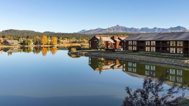 Una cabaña junto al lago con su reflejo en el agua, rodeada de montañas y follaje otoñal.