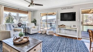 A spacious living room with a large TV, comfortable seating, a ceiling fan, and a view of the outdoors. - Surfside - Dock with bar area / Gentle lot / Cozy Retreat (Moneta)