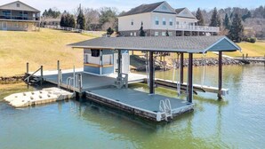 A dock with a covered seating area and a small building, likely a restroom, by a lake with houses in the background. - Surfside - Dock with bar area / Gentle lot / Cozy Retreat (Moneta)