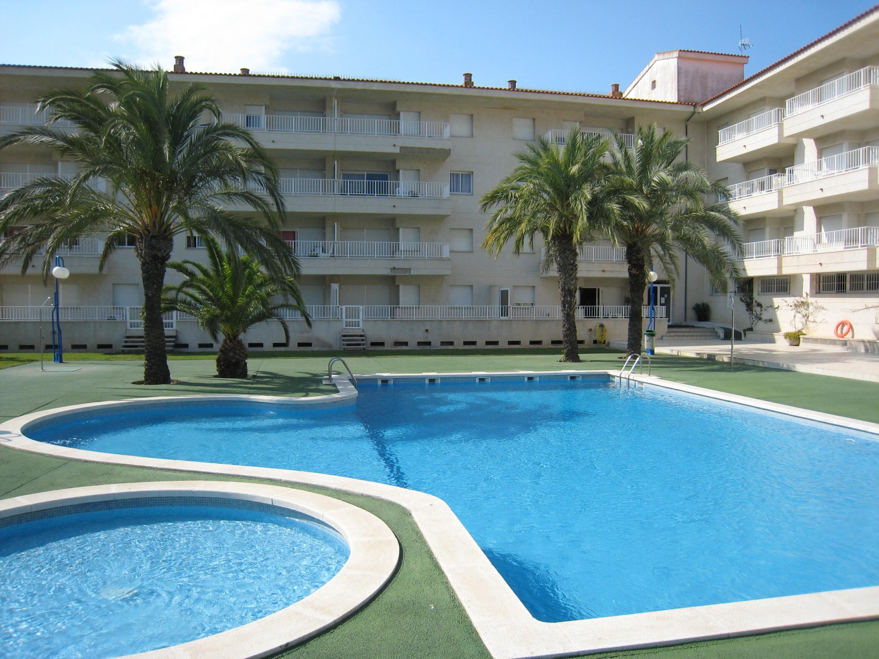 A swimming pool with a green border, surrounded by palm trees and a multi-story building with balconies.