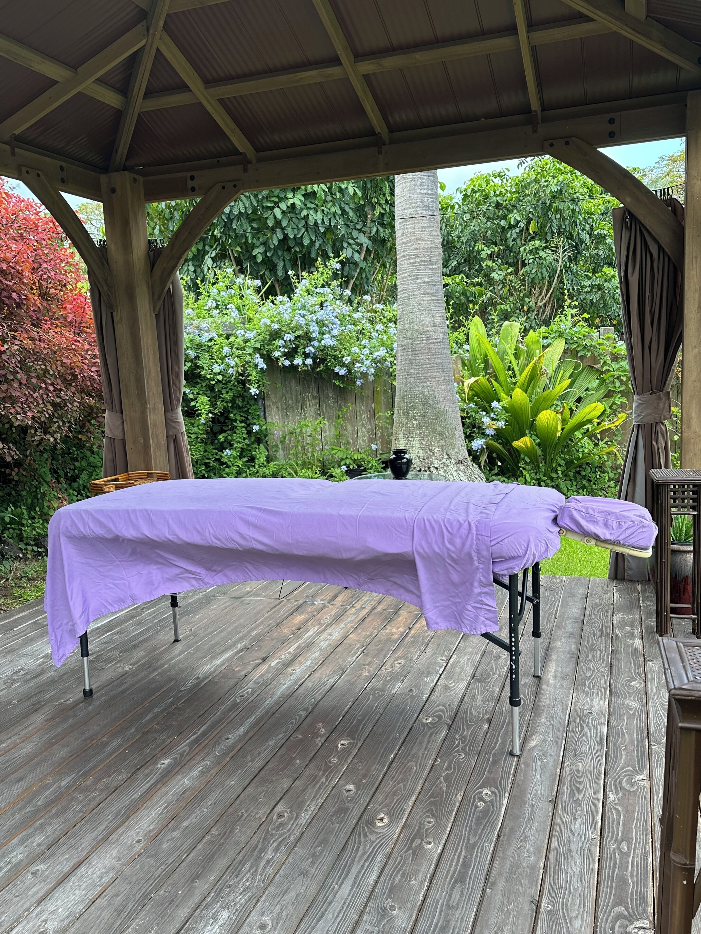 A covered outdoor area with a table covered in purple cloth, wooden deck flooring, and surrounding greenery.
