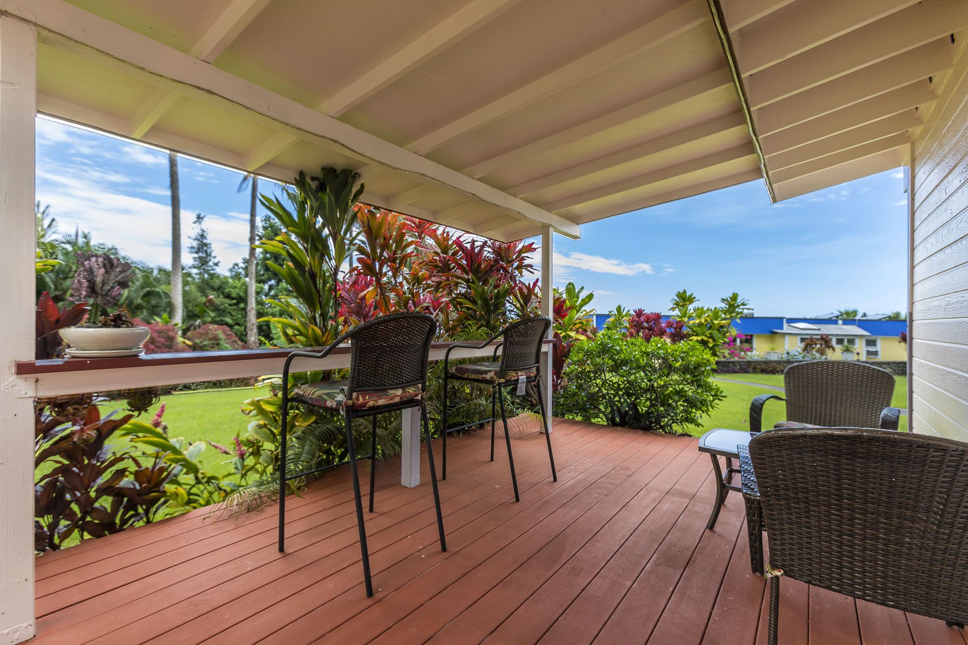 A covered outdoor patio with a wooden deck, two wicker chairs, a small table, and a potted plant.