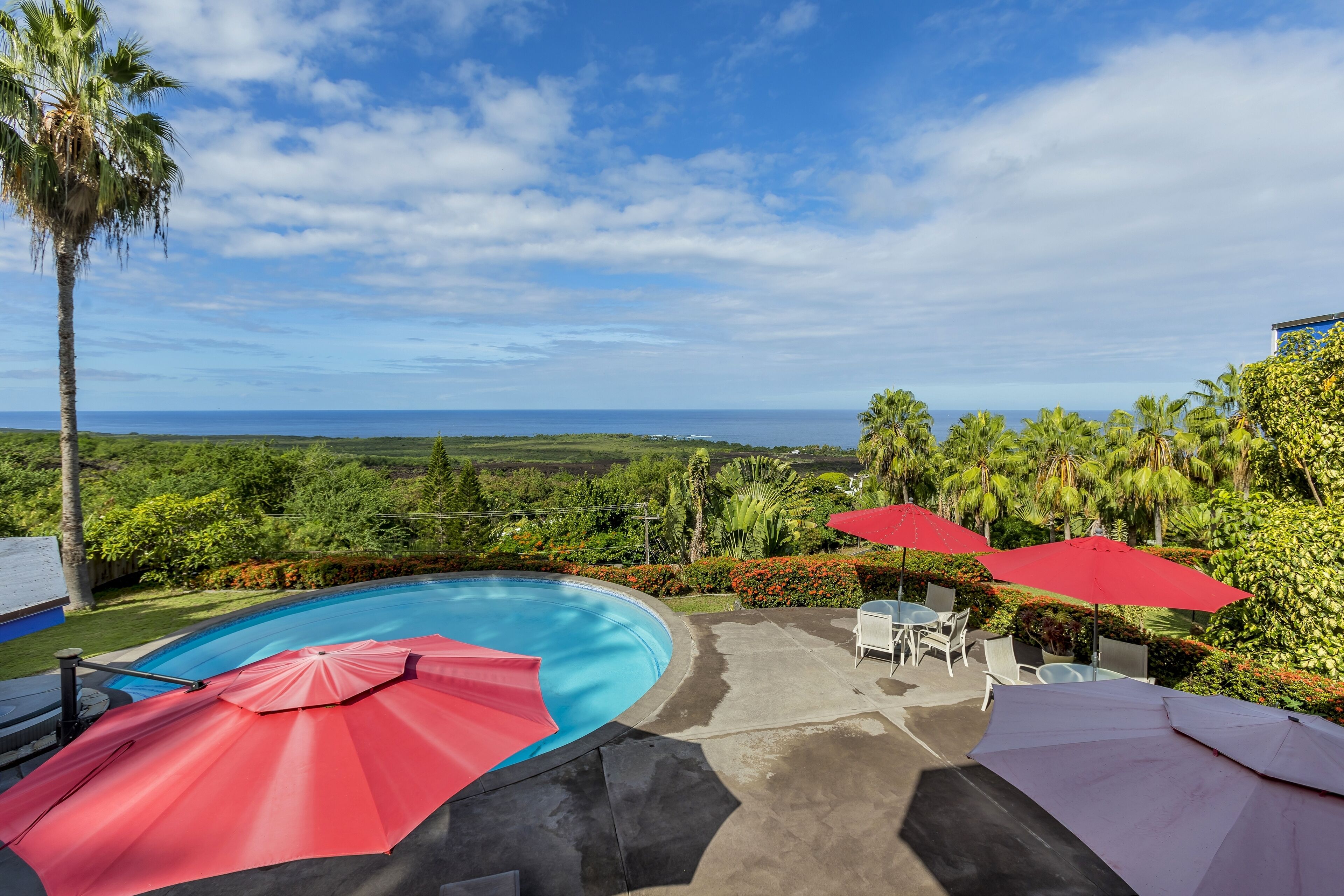 A pool area with red umbrellas, outdoor furniture, and a view of the ocean.