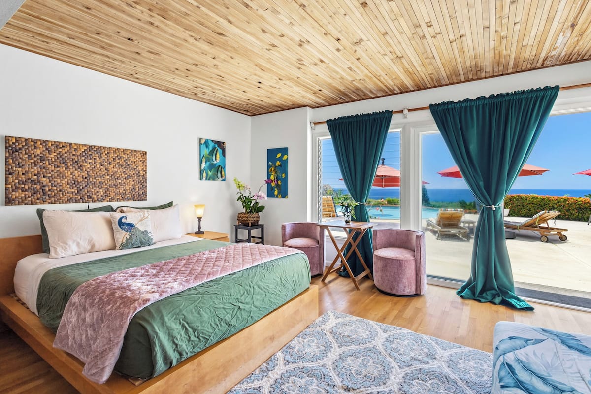 A bedroom with a wooden ceiling, a large bed, a view of the sea, and a balcony with chairs and umbrellas.