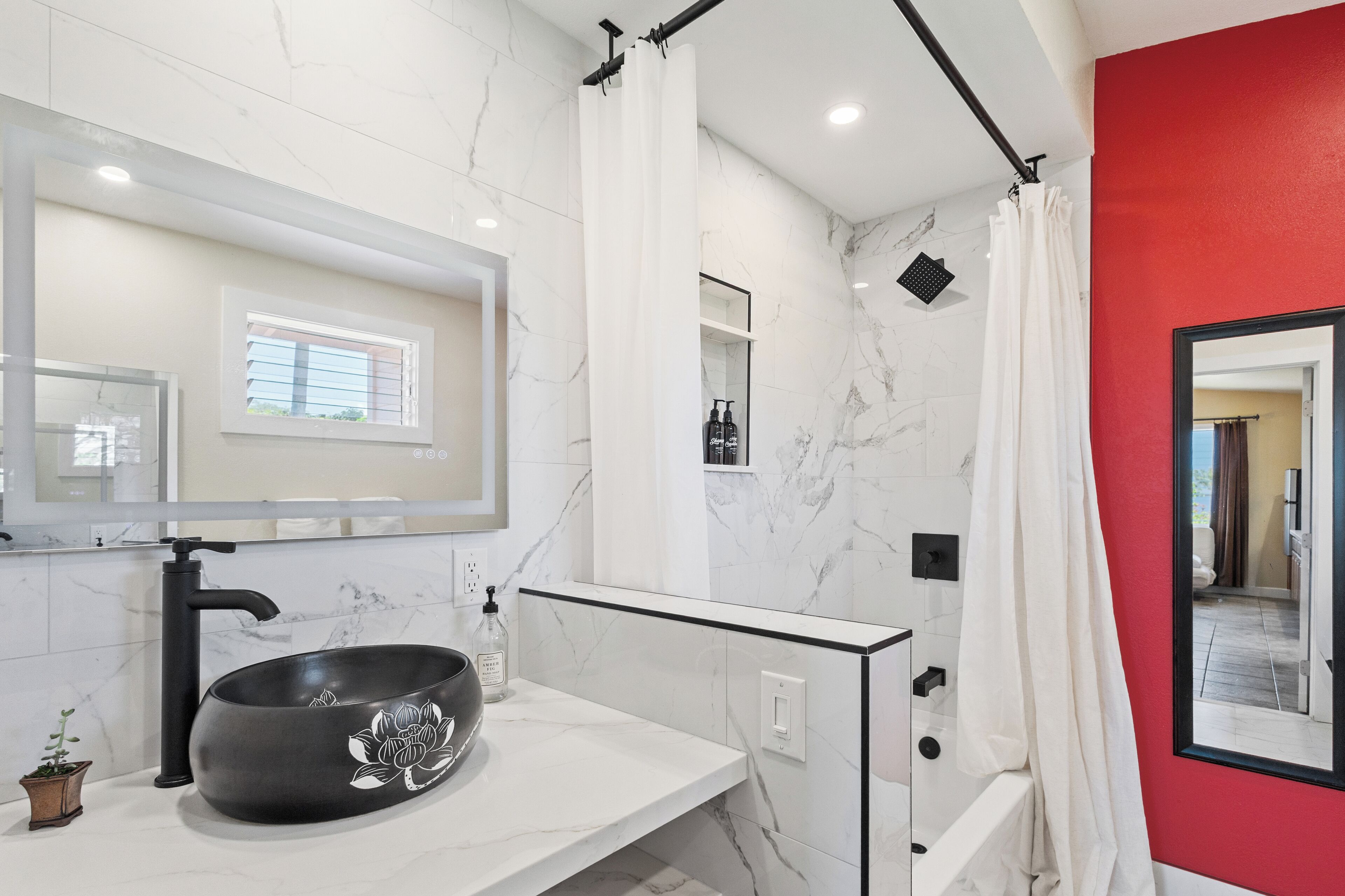 A bathroom with a large mirror, a black basin, a small plant, and a red accent wall.