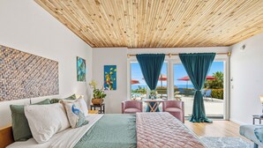 A bedroom with a wooden ceiling, a bed with green and white bedding, a view of the outdoors through a glass door, and a small table with chairs. - Luana Nai'a Inn (Captain Cook)