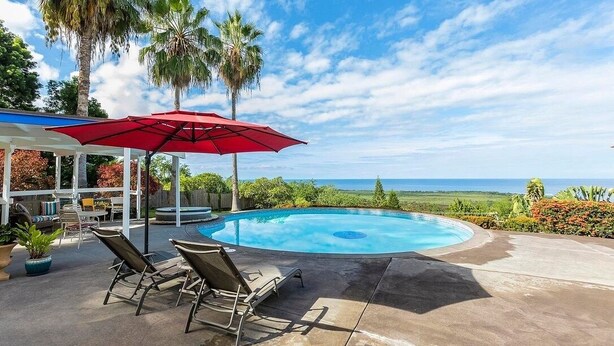 A pool area with lounge chairs and a red umbrella, surrounded by palm trees and a view of the ocean. - Luana Nai'a Inn (Captain Cook)