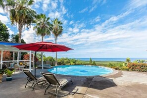 A pool area with lounge chairs and a red umbrella, surrounded by palm trees and a view of the ocean.