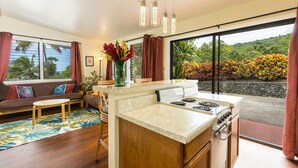 A modern kitchen with a central island, a dining area with a sofa and table, and large windows offering a view of greenery. - Luana Nai'a Inn (Captain Cook)