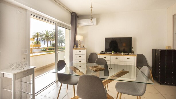 A modern living room with a glass dining table, grey chairs, a flat-screen TV, and a balcony with a view of palm trees. - Apartment with swimming pool, close to the sea (Roses)