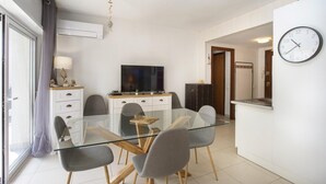 A modern living room with a glass dining table, grey chairs, a flat-screen TV, and a clock on the wall. - Apartment with swimming pool, close to the sea (Roses)