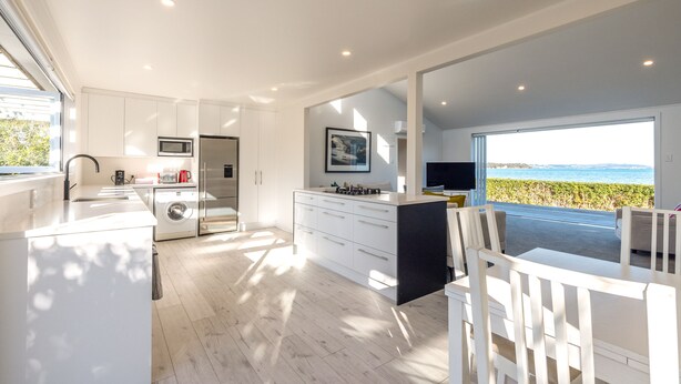 A modern kitchen with white cabinetry, a built-in microwave, and a large window overlooking a scenic view. - Driftwood Beach House with Spa Pool (Waiheke Island)