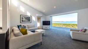 A modern living room with a sofa, coffee table, and TV, featuring a large window with a view of the sea. - Driftwood Beach House with Spa Pool (Waiheke Island)