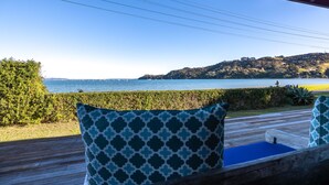 A wooden deck with two patterned cushions, overlooking a body of water and a hilly landscape. - Driftwood Beach House with Spa Pool (Waiheke Island)