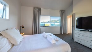 A hotel room with a large bed, a TV, a window with curtains, and a view of the sea. - Driftwood Beach House with Spa Pool (Waiheke Island)